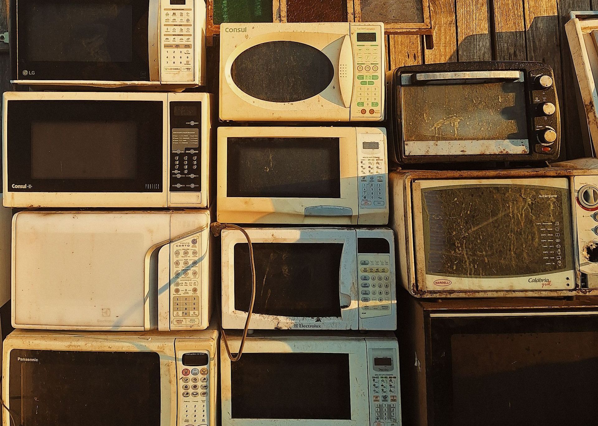 A stack of old microwaves placed outdoors against a rustic wooden wall in warm sunlight.